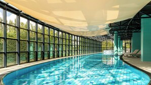 Una amplia piscina interior con baldosas azules brillantes, rodeada de grandes ventanales que ofrecen vistas a zonas verdes.
