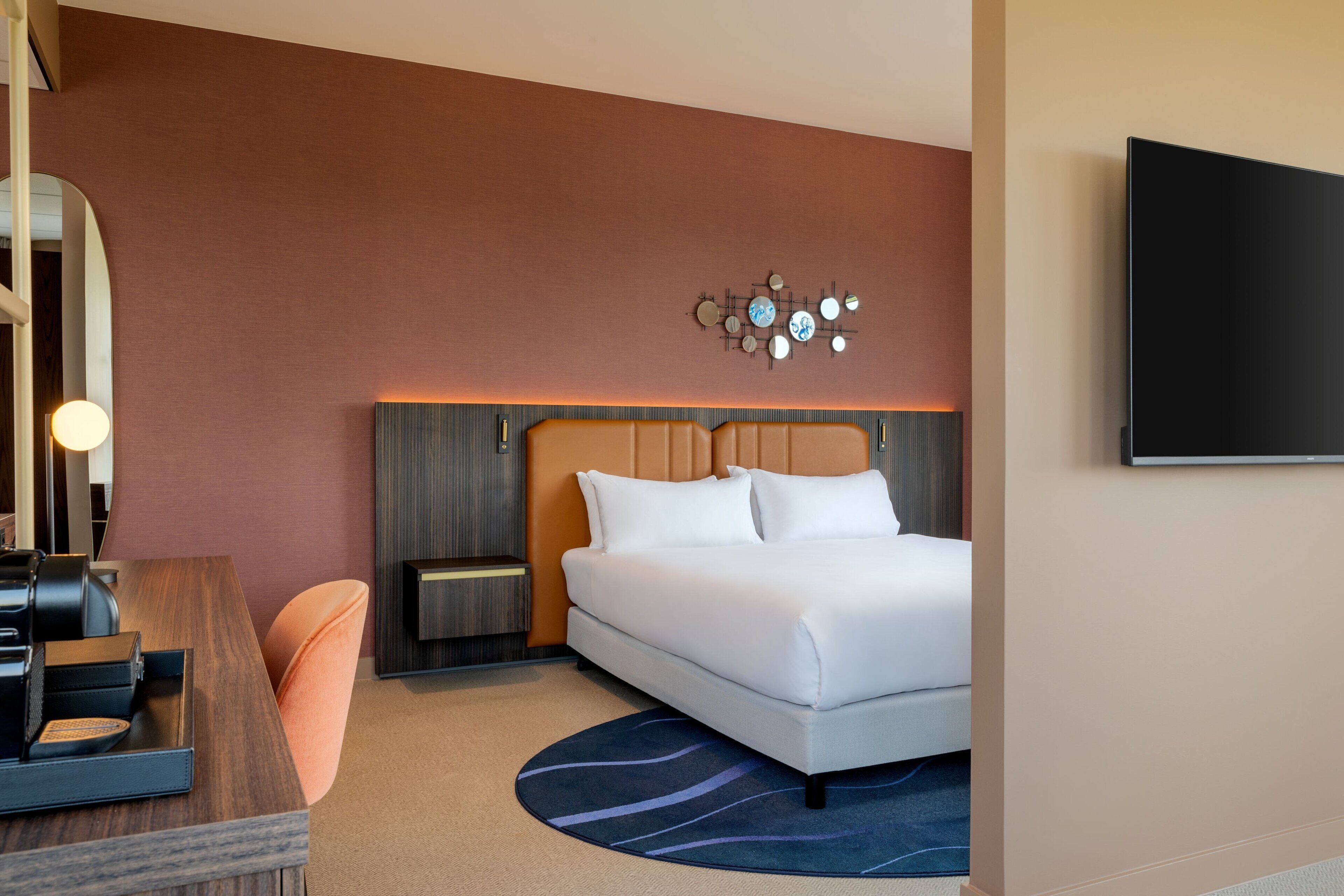 Une chambre d’hôtel moderne comprenant un lit, un bureau avec une chaise, une table de chevet, un téléviseur fixé au mur et un élément décoratif mural.