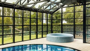 Una piscina interior moderna con techo de vidrio, un jacuzzi y vistas a un jardín cuidado.