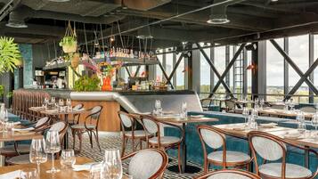 A restaurant with multiple dining tables and chairs, a bar area, and large windows offering a view.