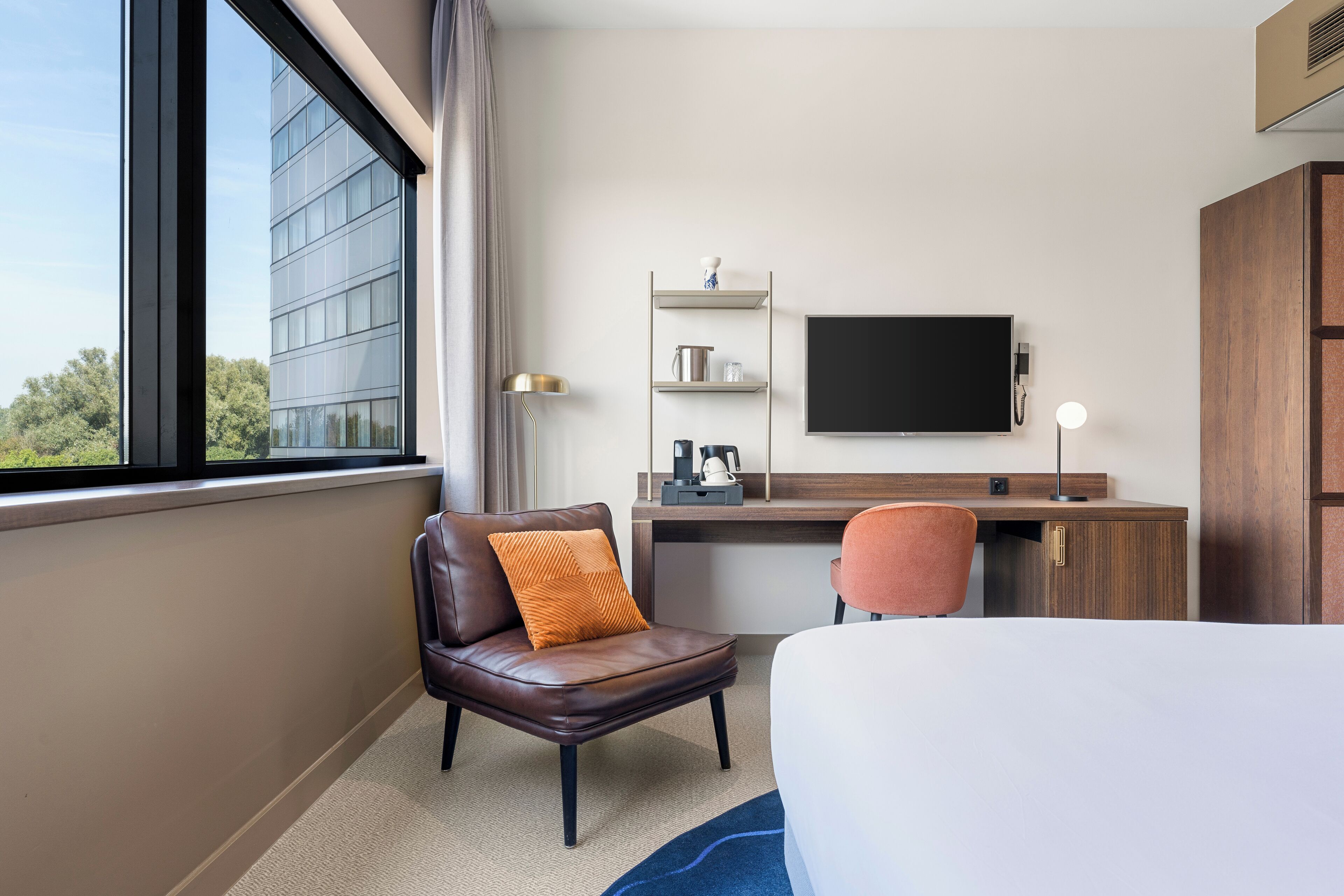 A hotel room with a bed, a desk with a flat-screen TV, a chair, a small shelf with items, and a window with a view of a building.