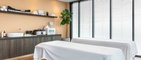 A modern spa room with two treatment tables, a reception desk, and a large window with blinds.