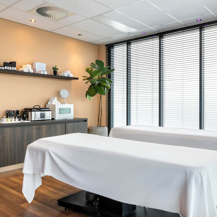 A modern spa room with two treatment tables, a reception desk, and a large window with blinds.