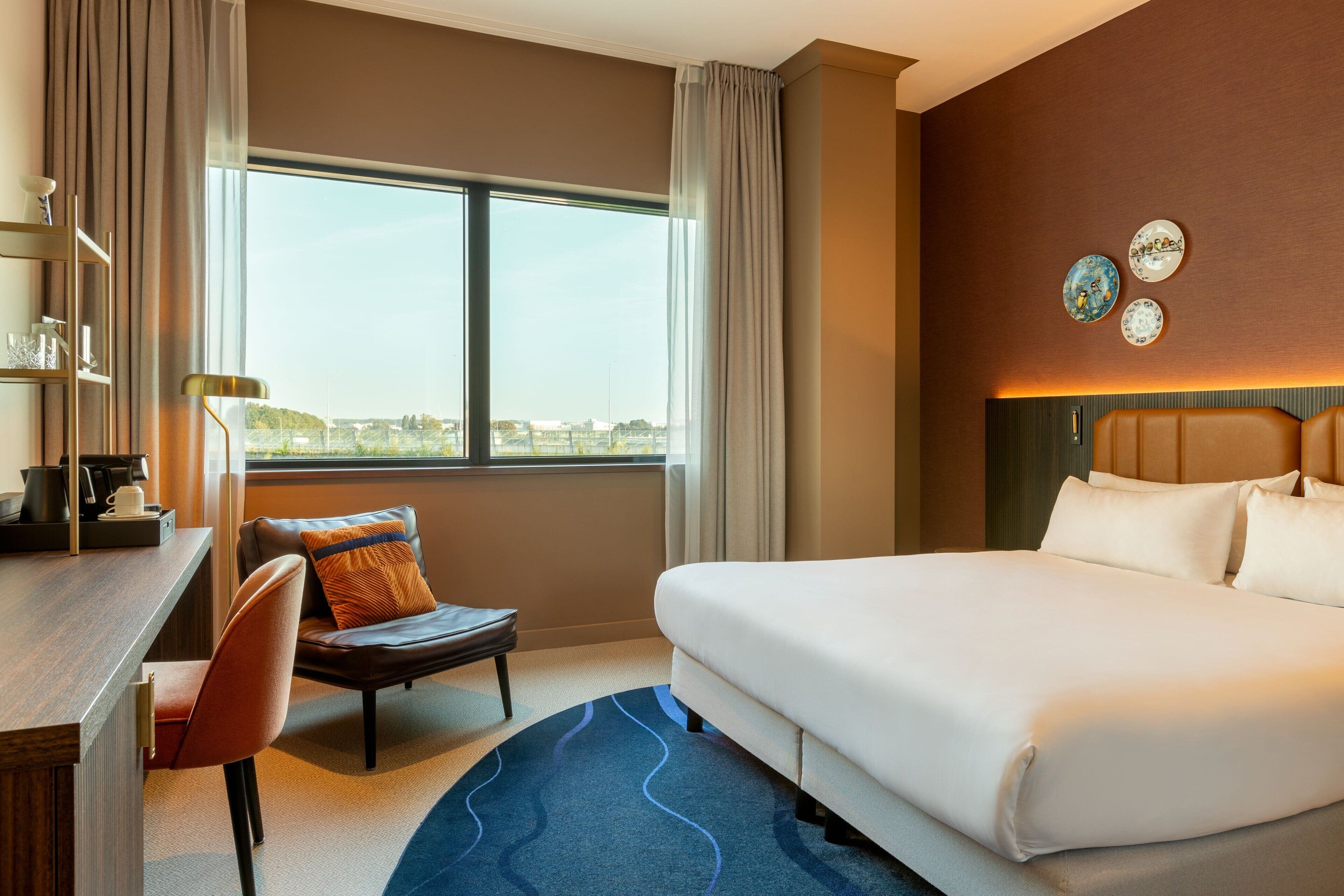 Habitación de hotel con una cama grande, un escritorio con silla, vista a la ciudad y platos decorativos en la pared.