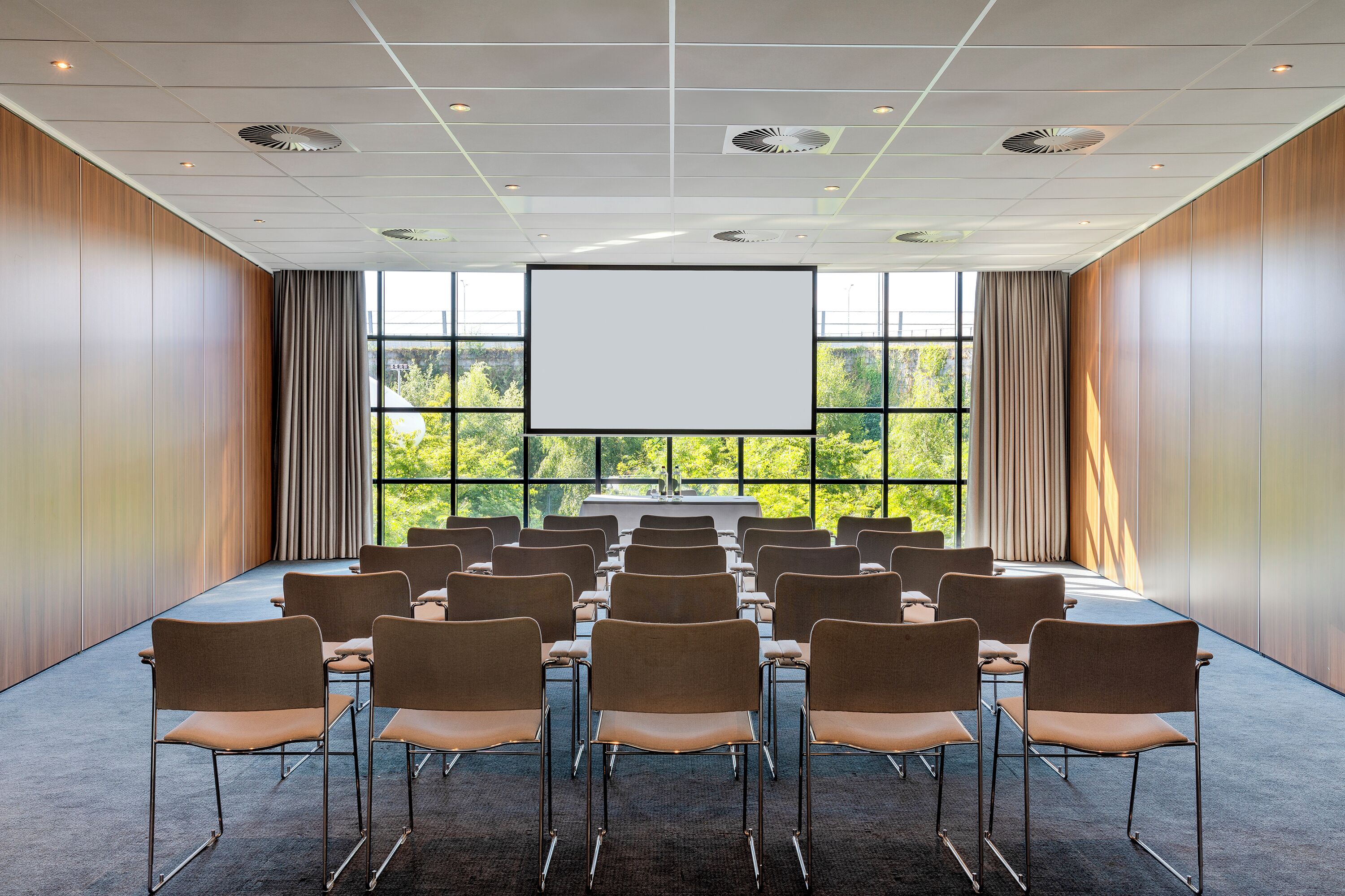 A conference room with rows of chairs facing a large screen, wooden panel walls, and large windows with a view of greenery.