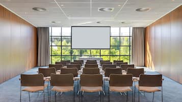 A conference room with rows of chairs facing a large screen, wooden panel walls, and large windows with a view of greenery.