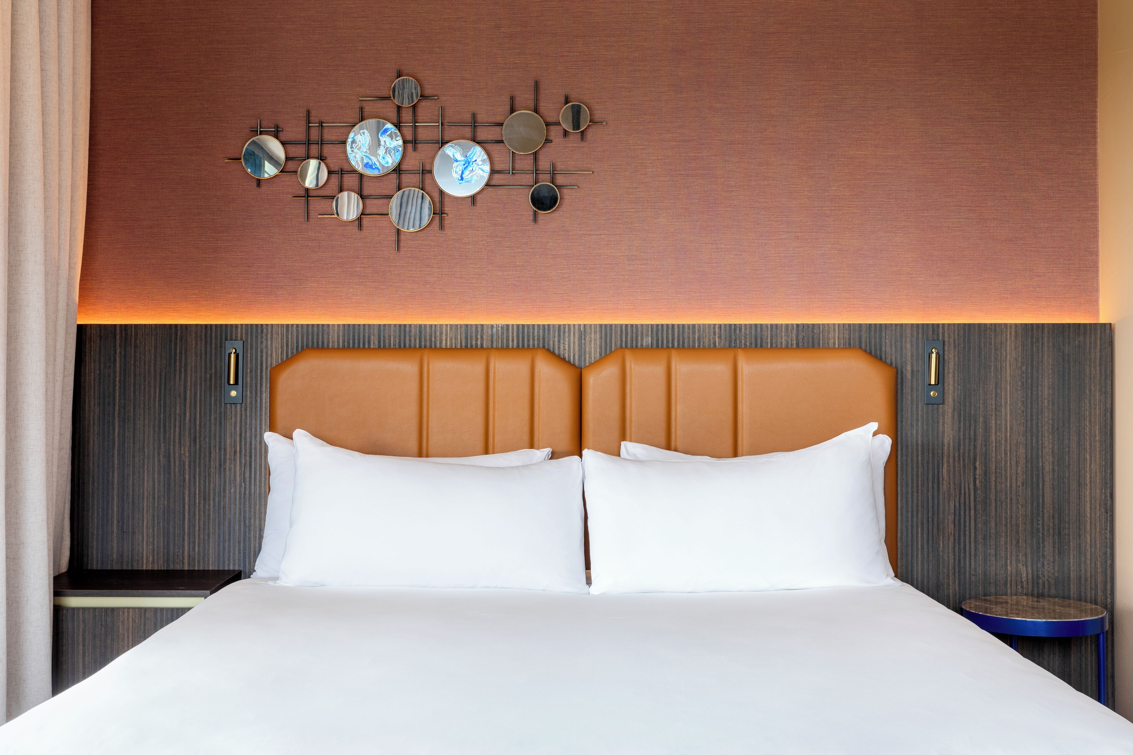 A hotel room with a large bed, two pillows, a headboard, a wall-mounted clock, and a small table.