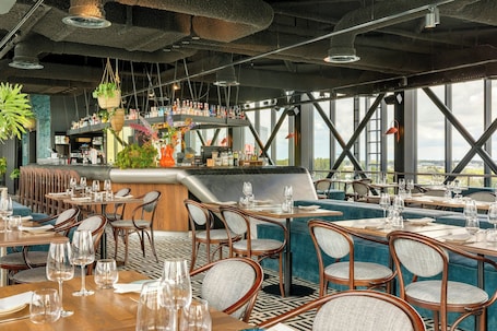 A restaurant with high ceilings, hanging plants, and a bar area. Tables are set with chairs and wine glasses.