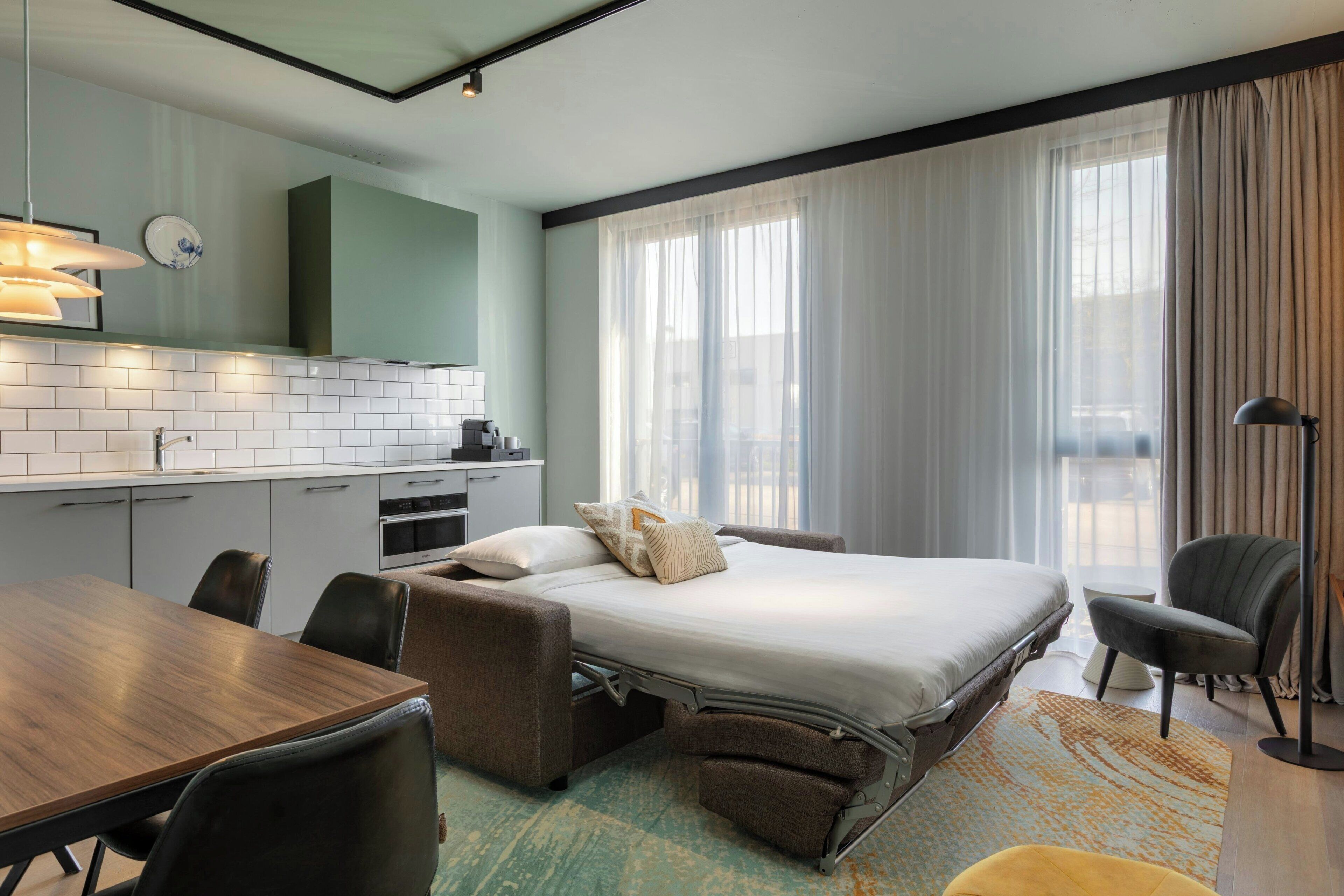 A hotel room with a bed, bedside table, chairs, a desk, a kitchenette, and large windows with curtains.
