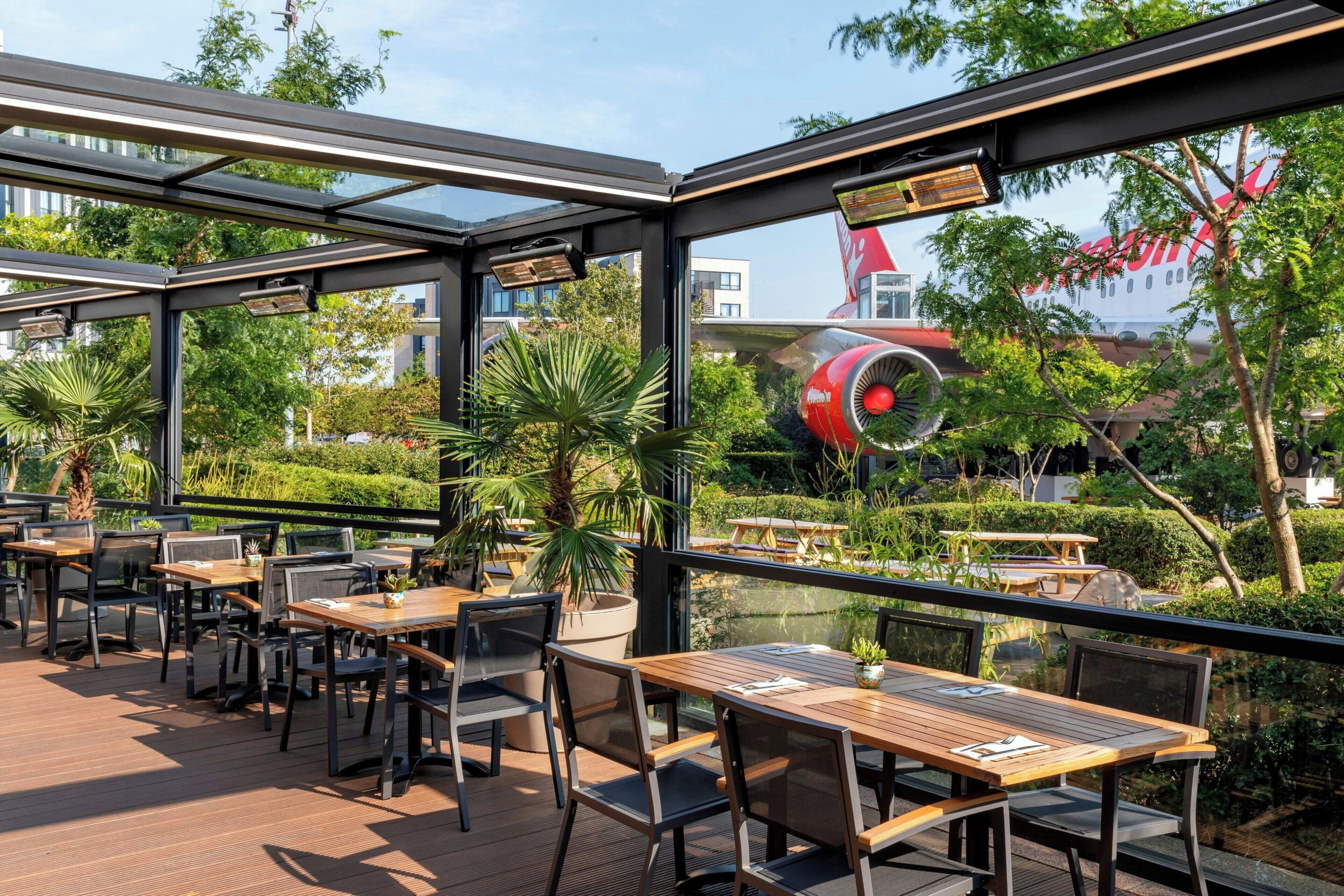 An outdoor dining area with tables and chairs, a glass enclosure, and a view of a red airplane.