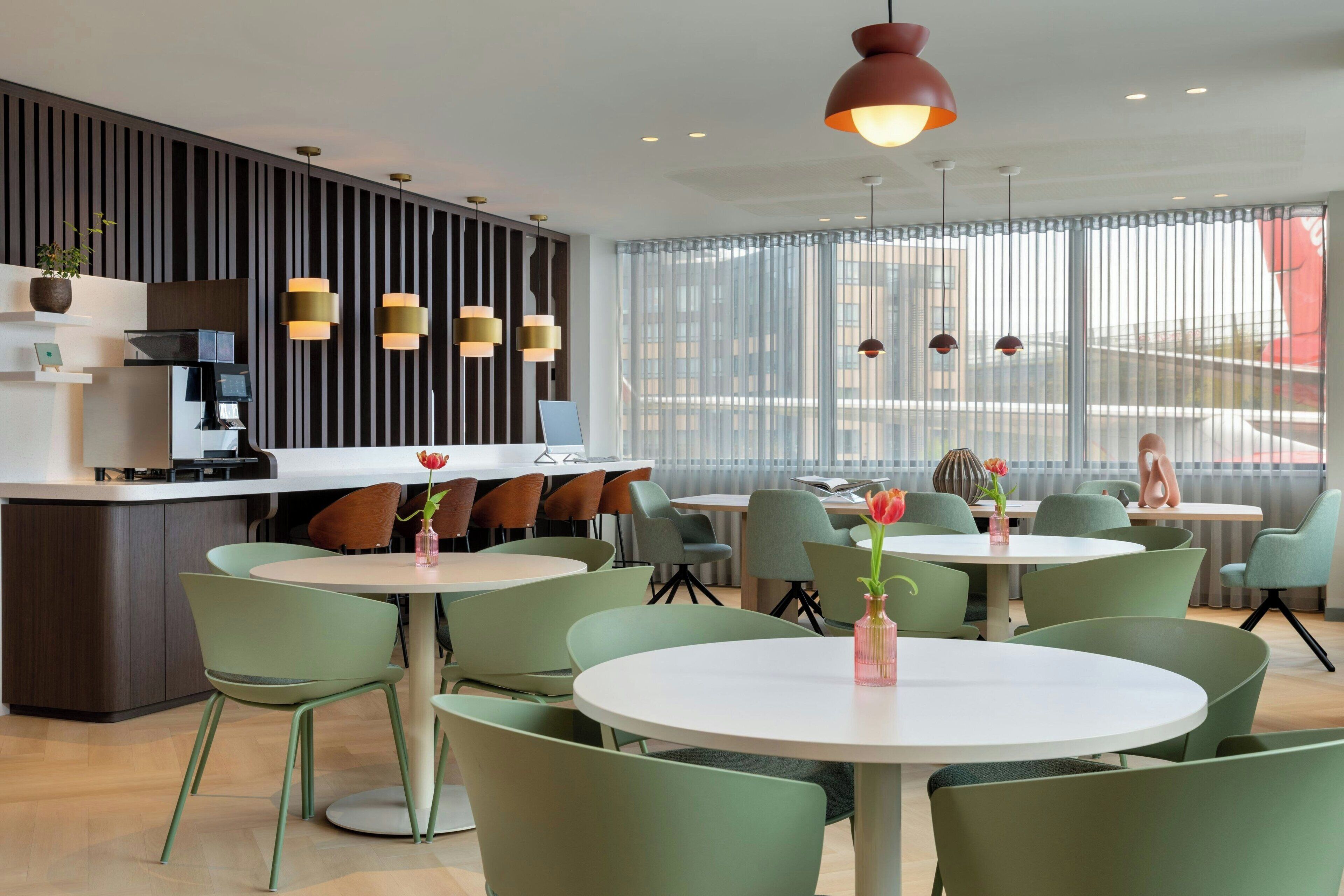 A modern restaurant with round tables and green chairs, a bar area, and large windows with a view.