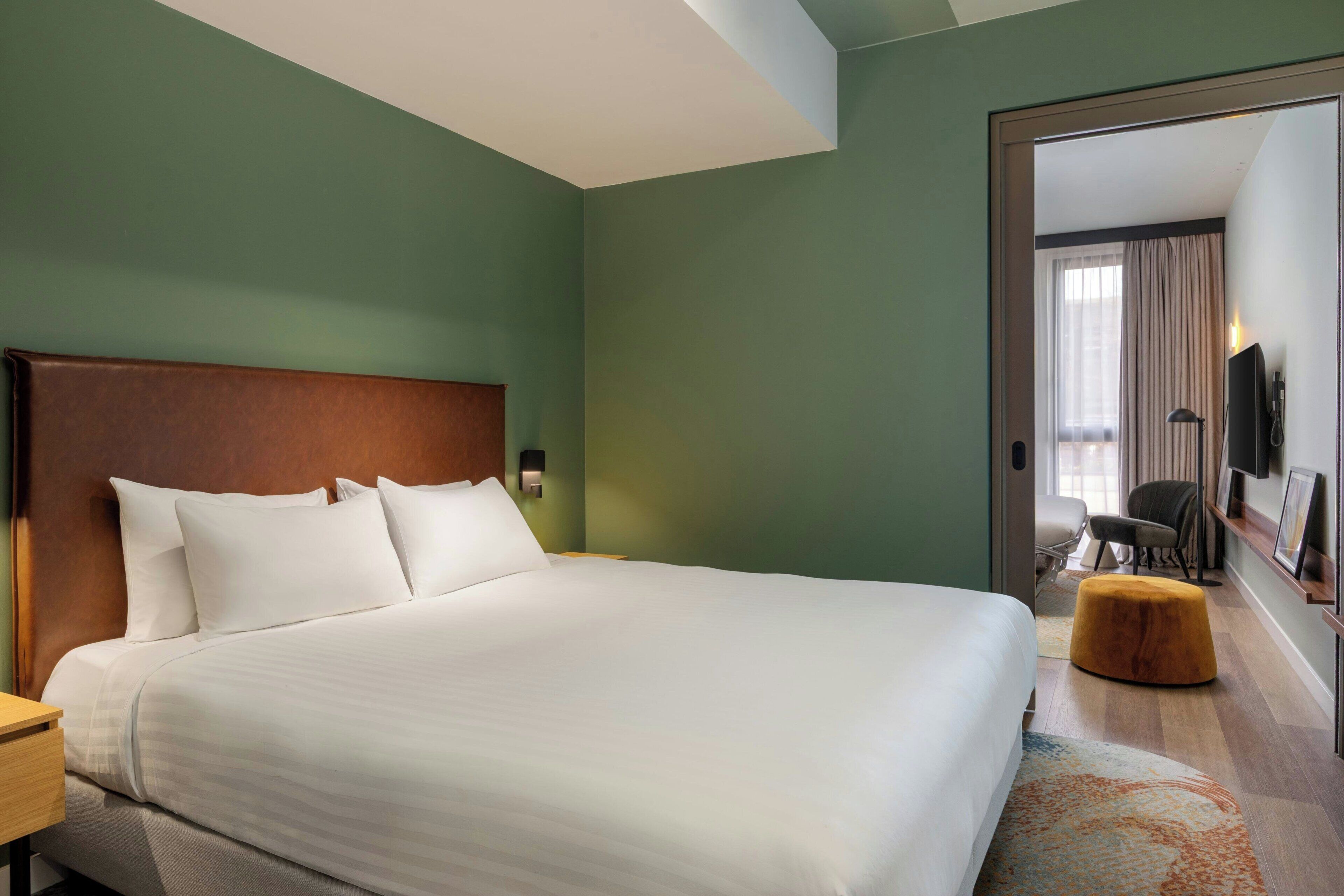 Una camera d'albergo con un letto grande, una testiera in legno, un poggiapiedi, un comodino, una scrivania, una sedia, una televisione e uno specchio.