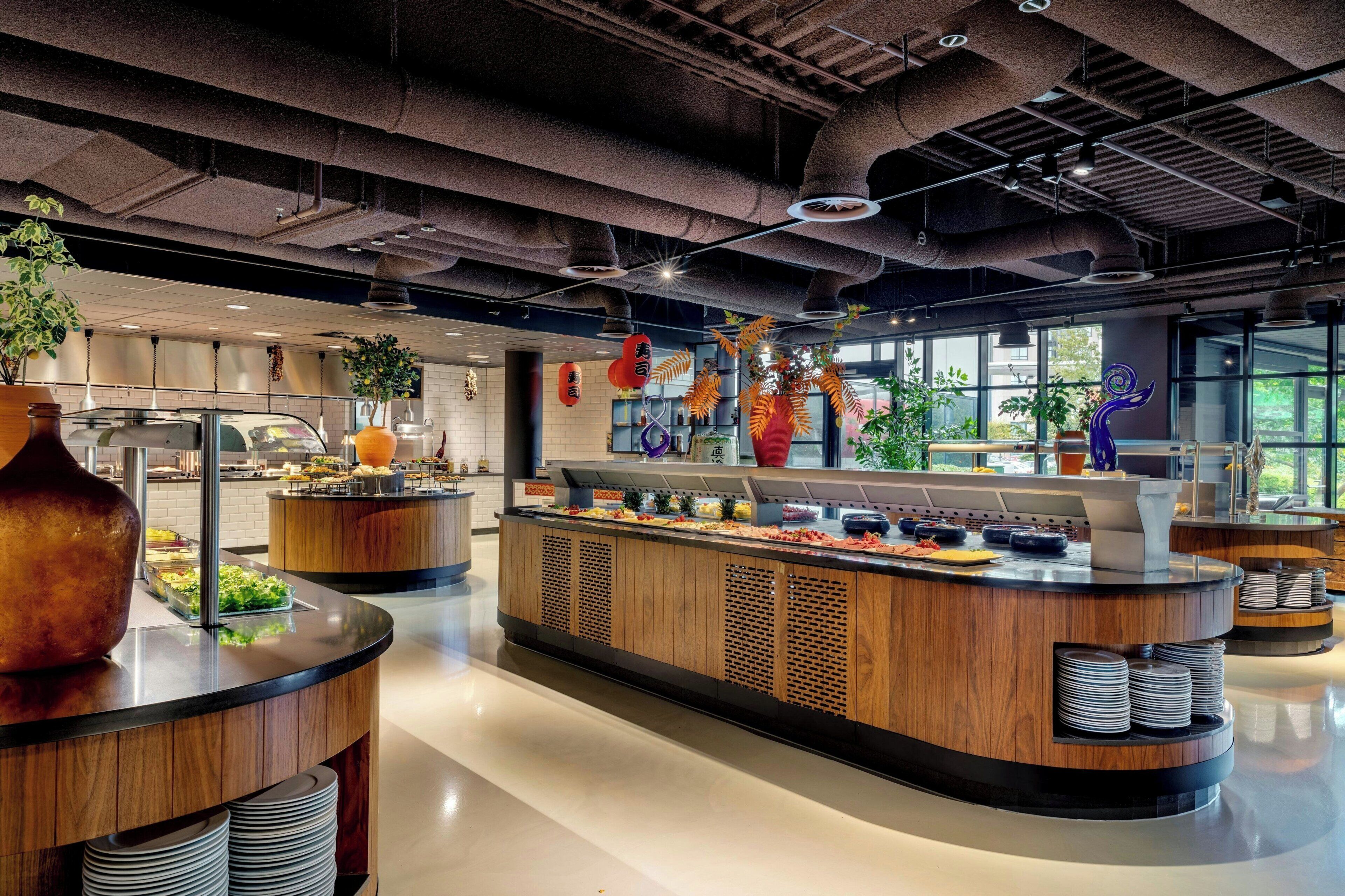 A modern restaurant with a central buffet, wooden accents, and large windows.