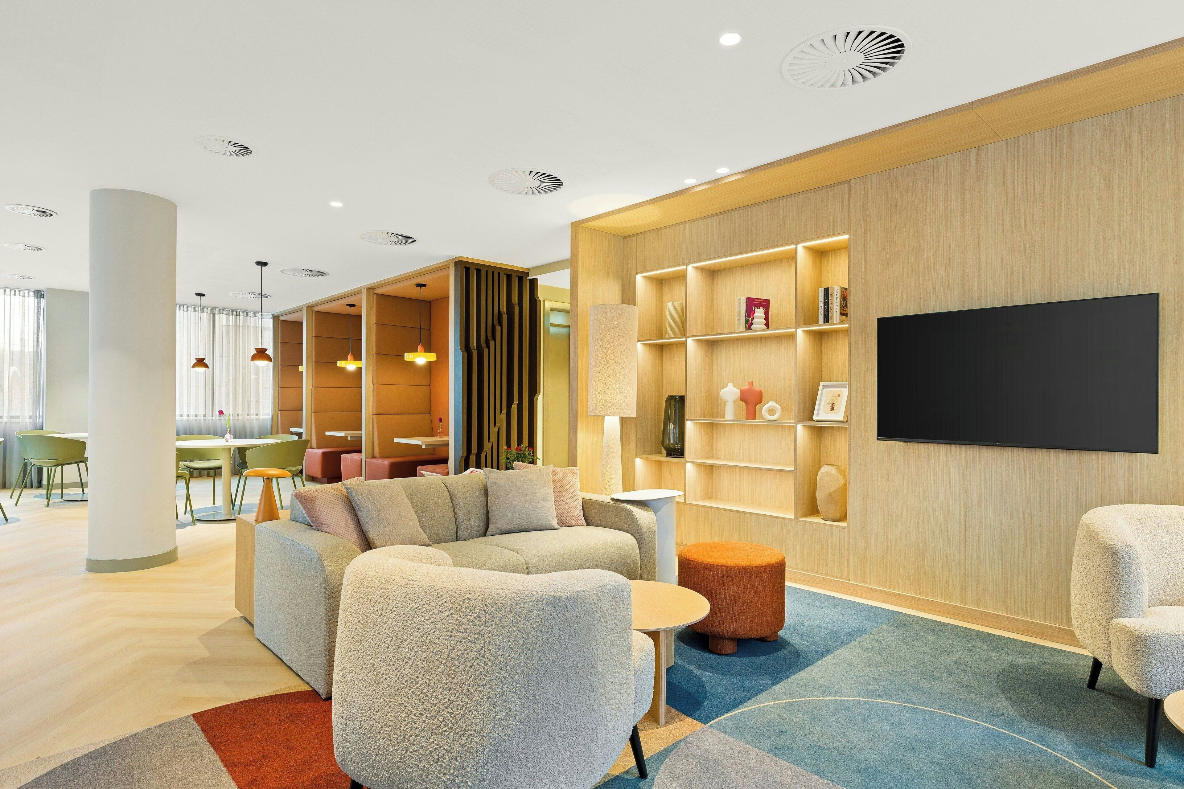 A modern hotel lobby with a sofa, ottoman, and a flat-screen TV mounted on the wall.