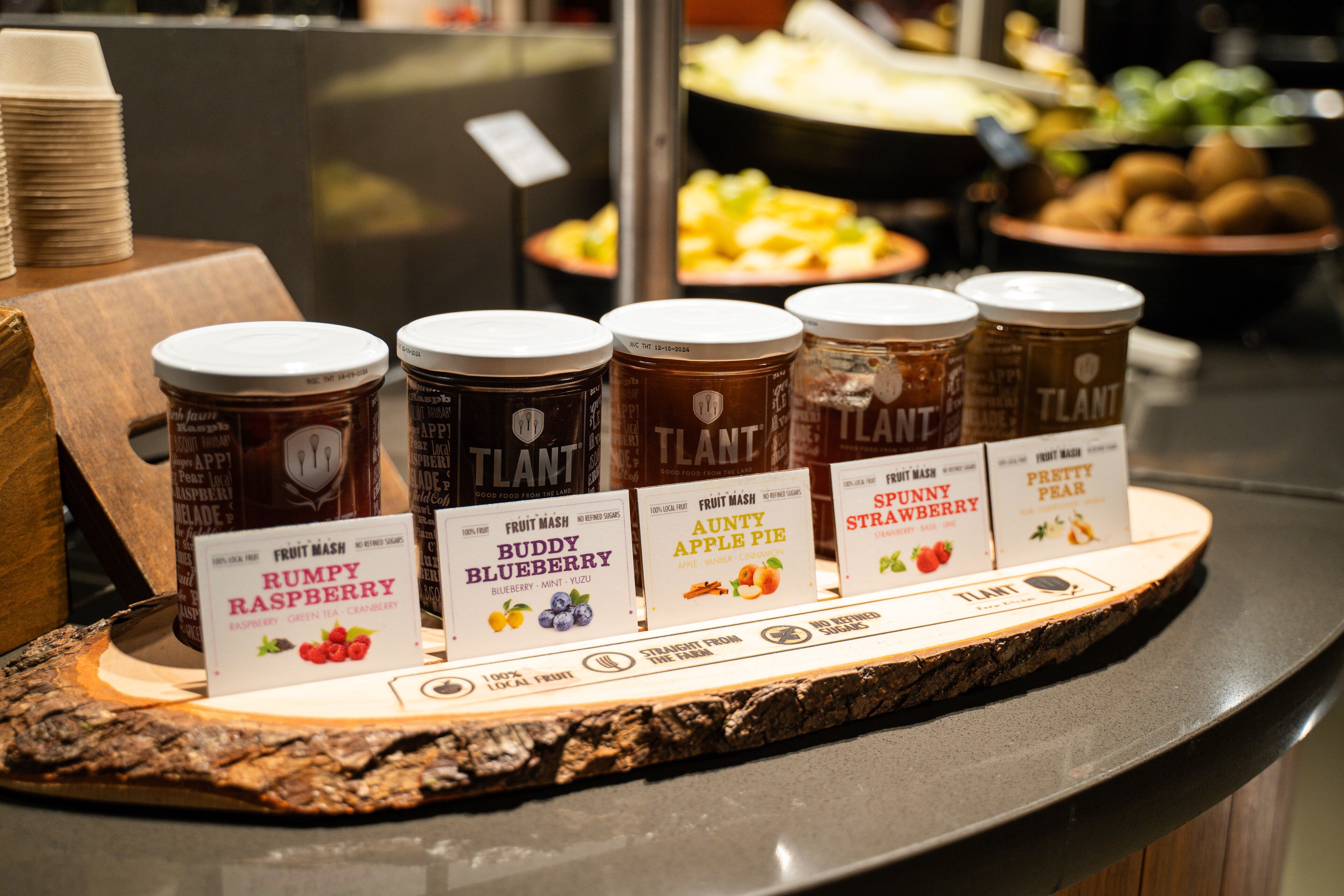 A display of fruit preserves with labels, including Rumpy Raspberry, Buddy Blueberry, Auntie Apple Pie, and others, on a wooden board.