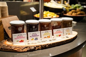 A display of fruit preserves with labels, including Rumpy Raspberry, Buddy Blueberry, Auntie Apple Pie, and others, on a wooden board.
