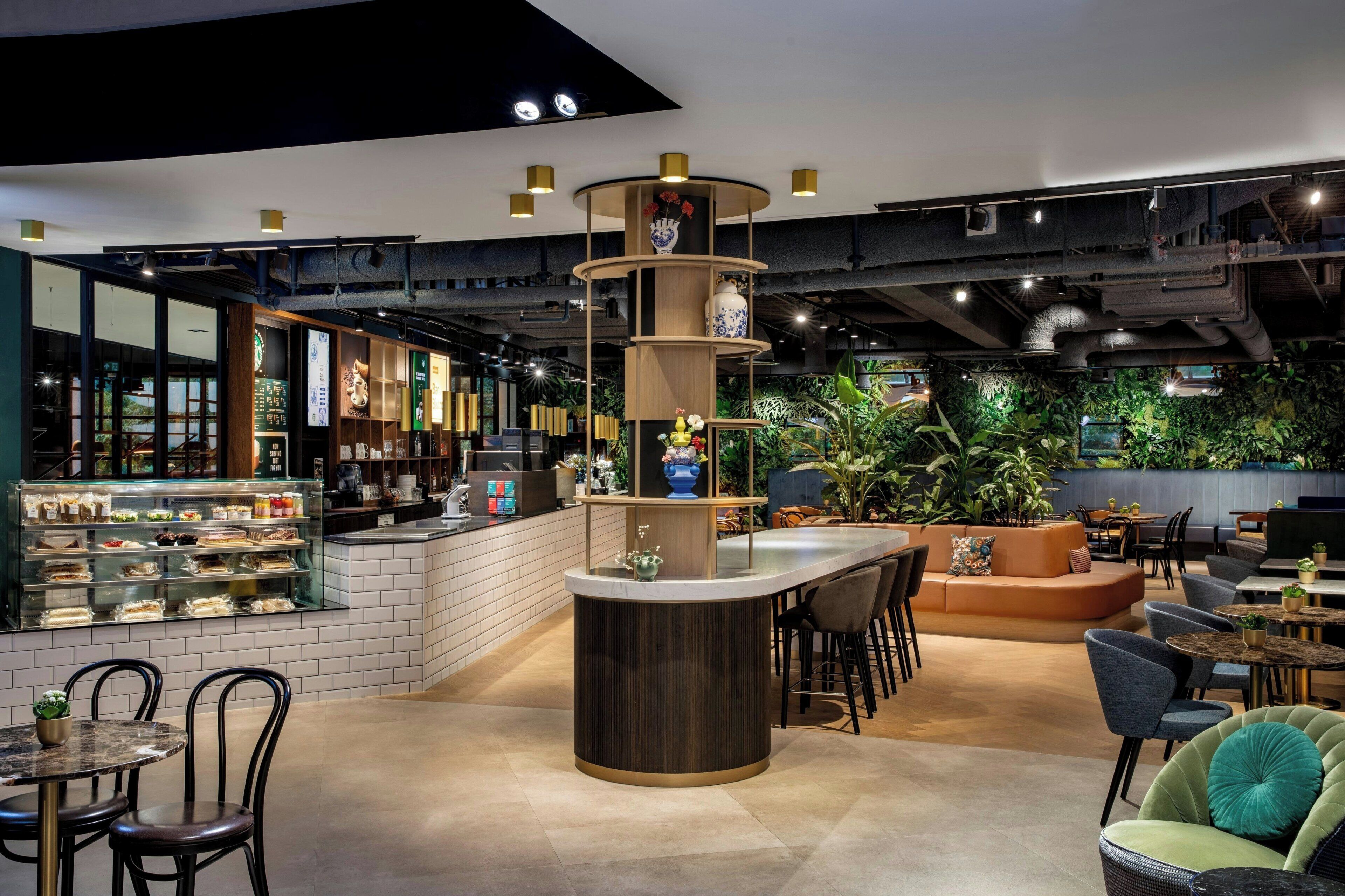 Een modern café met een centrale bar, zitgedeeltes en een groene wand met planten.