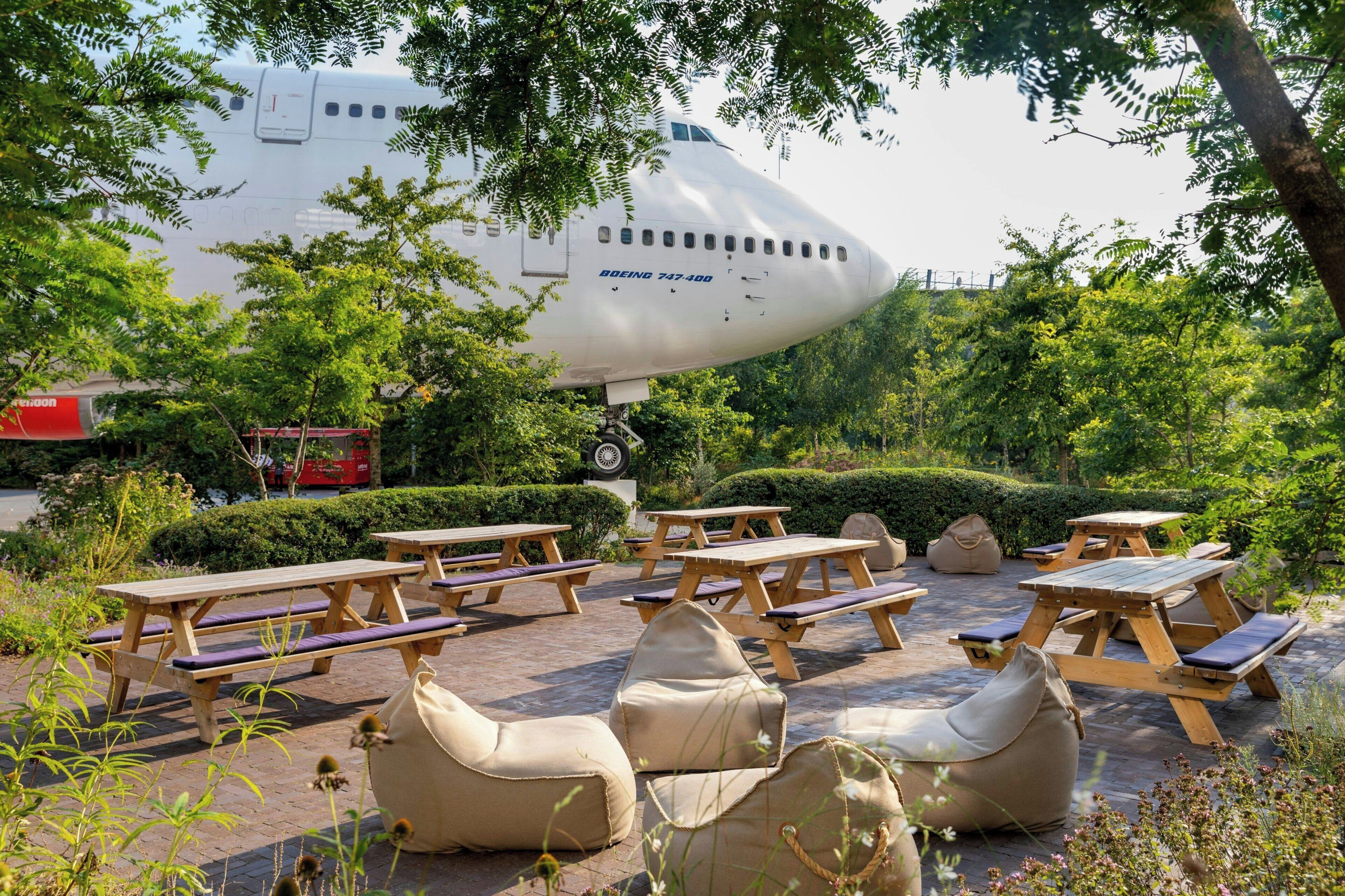 A garden area with picnic tables and bean bags, an airplane in the background, and a clear sky.