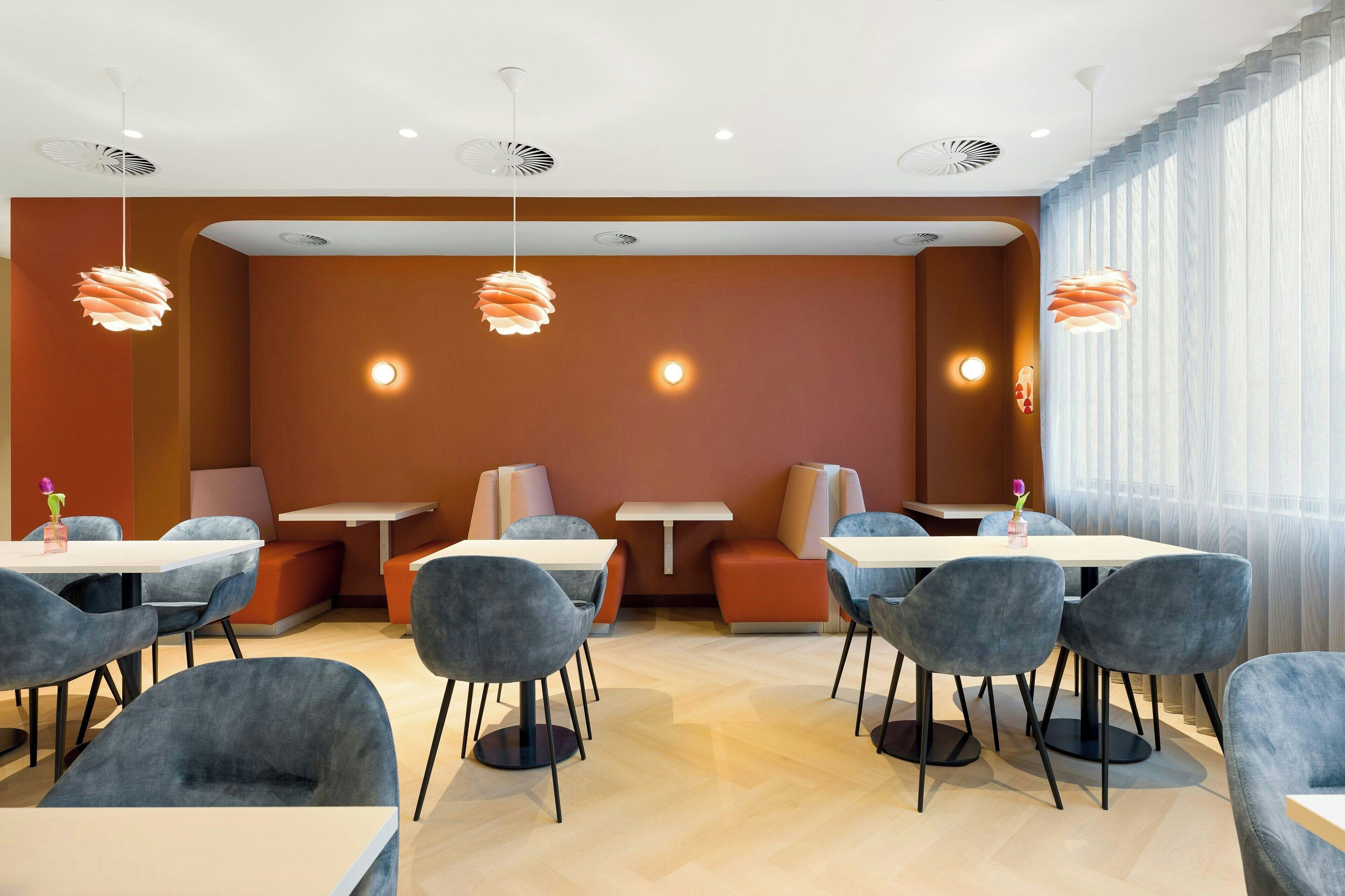 Zona pranzo moderna con tavoli rotondi e sedie grigie, posti a sedere arancioni e illuminazione a sospensione.