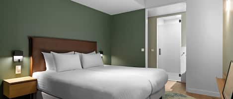 A modern hotel room with a large bed, a nightstand, a wall-mounted lamp, and a door leading to another room.