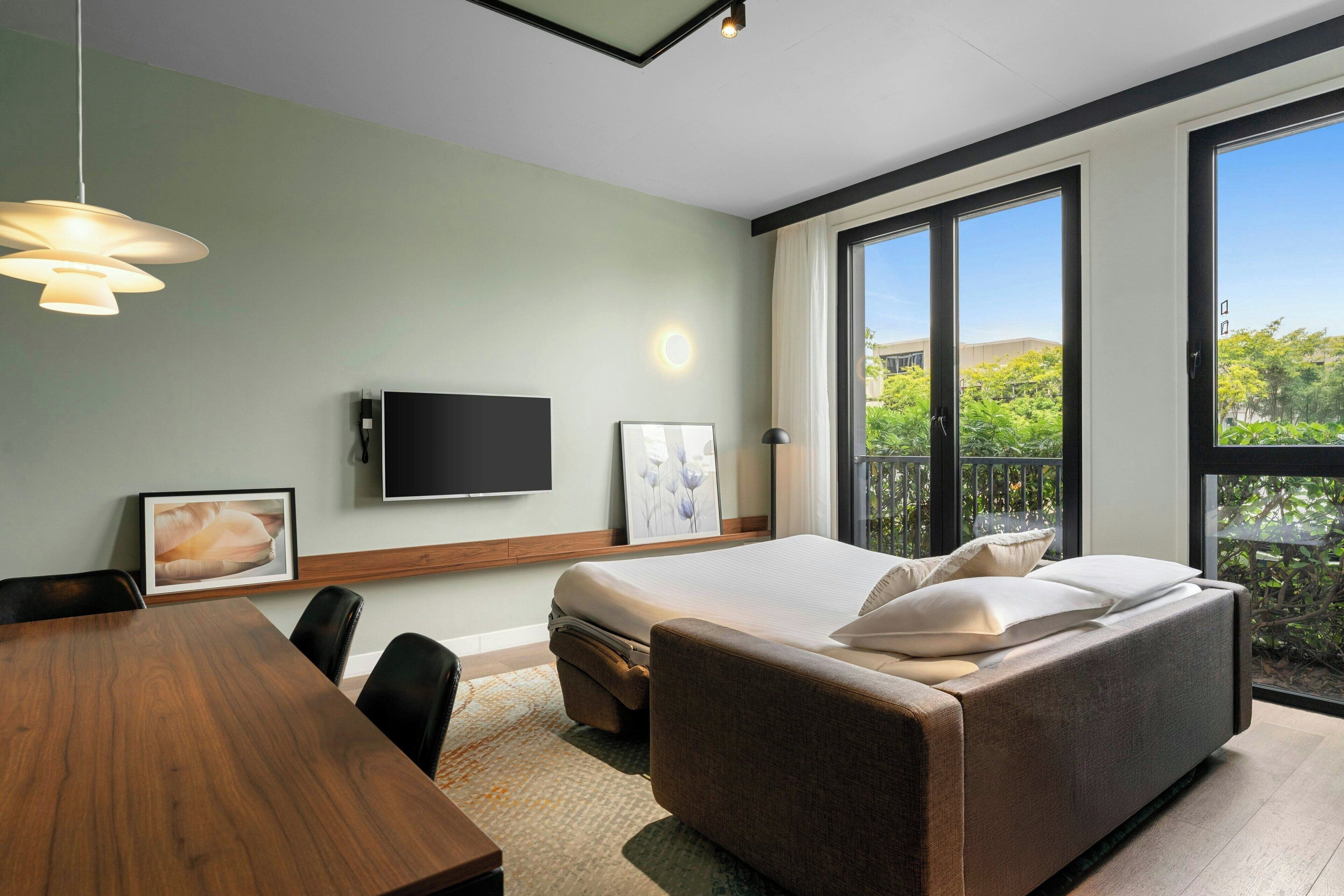 A modern hotel room with a large bed, a dining table, and a TV mounted on the wall.