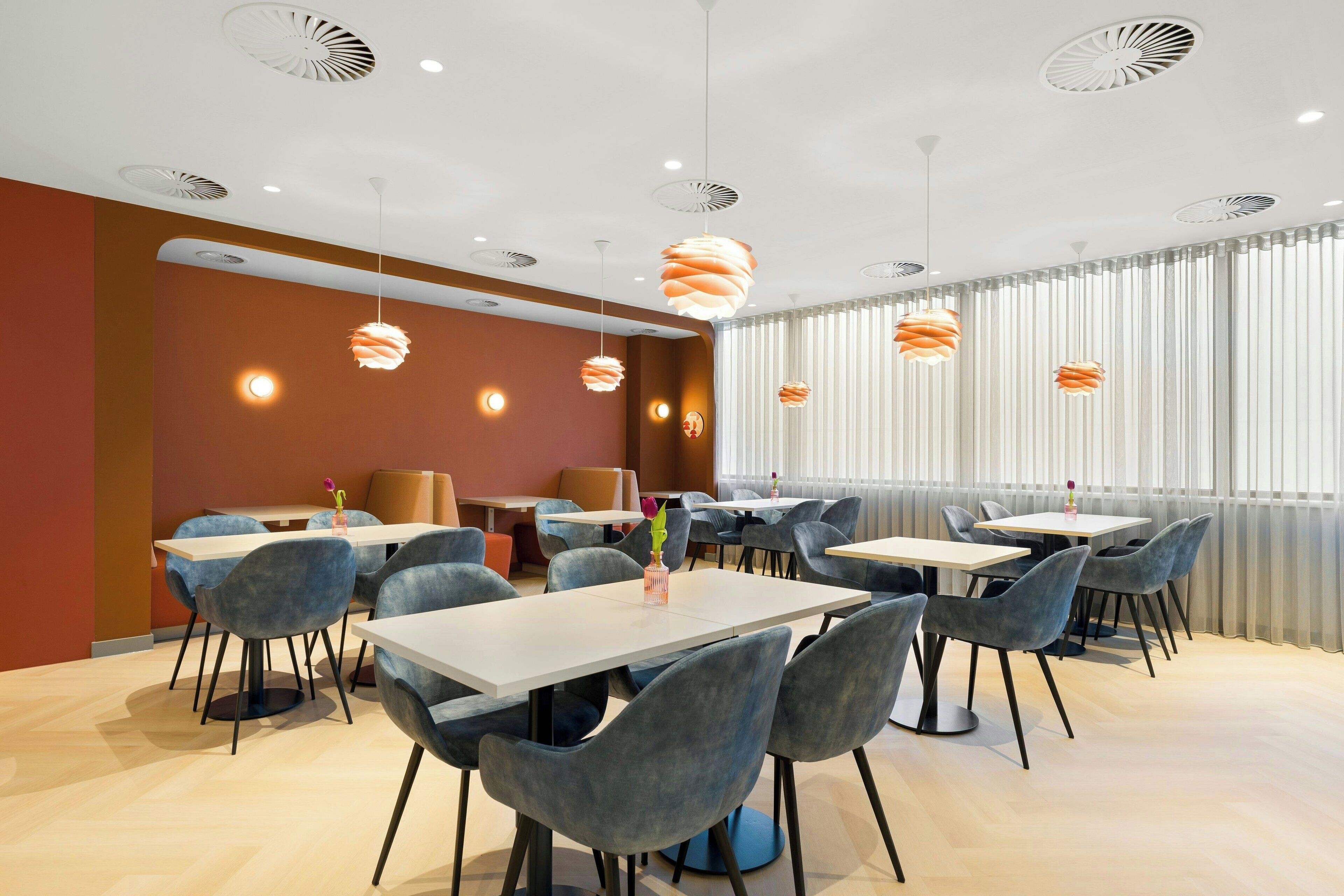 A modern dining area with round tables and chairs, ceiling fans, and a large window with blinds.