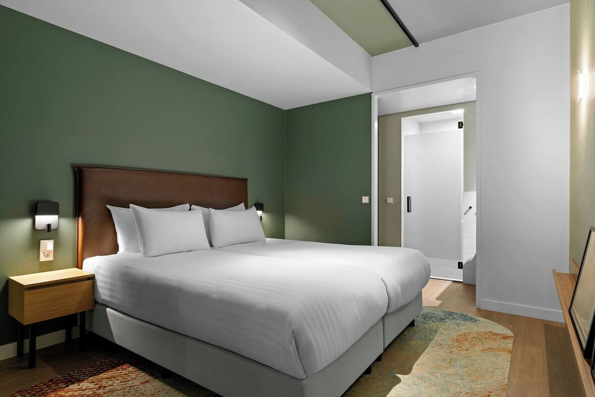 A modern hotel room with a large bed, a nightstand, a wall-mounted lamp, and a door leading to another room.