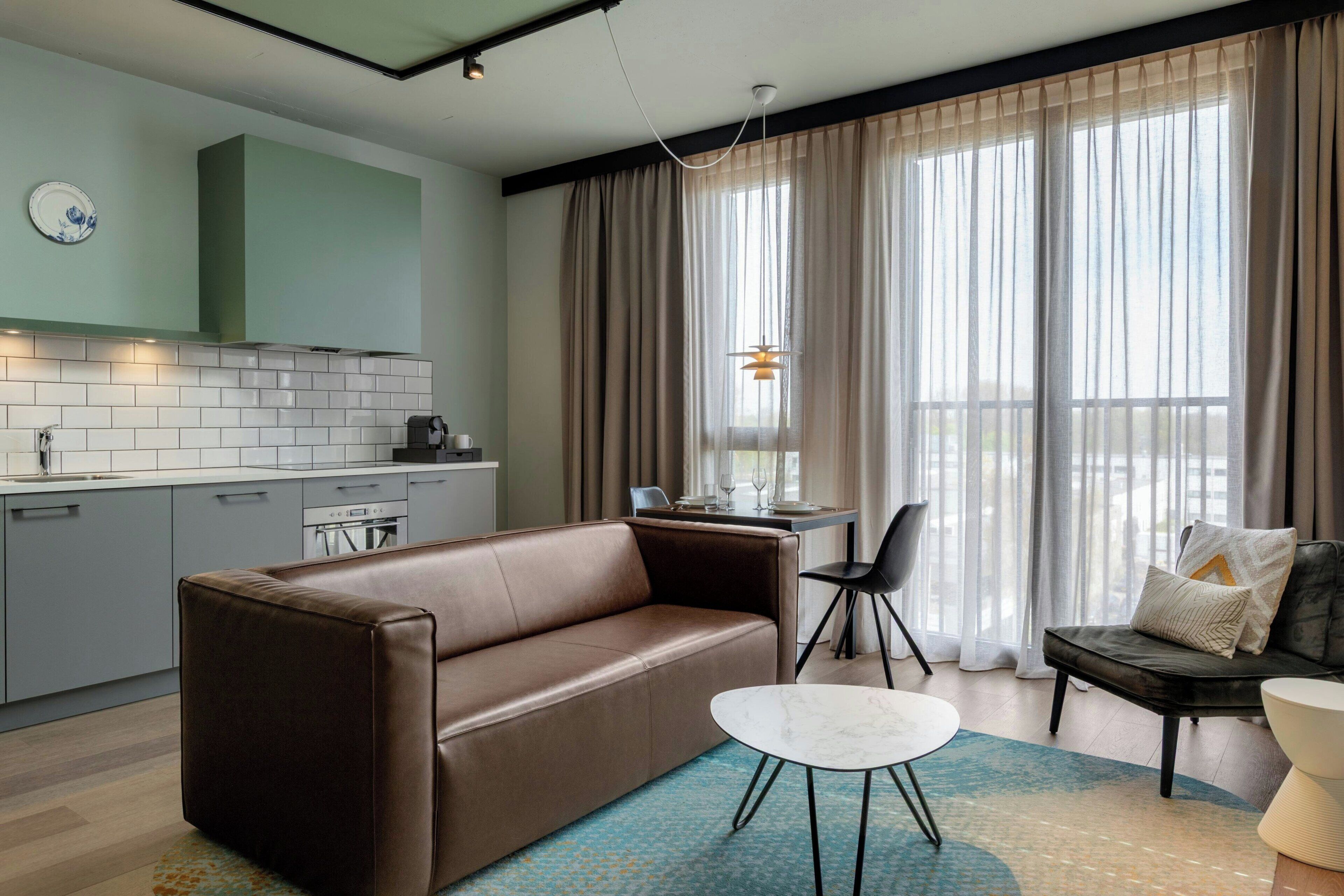 A modern living room with a brown leather sofa, a small white table, and a dining area with a table set for two.