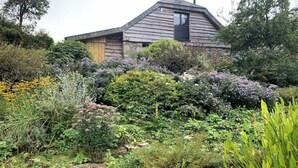 A rustic house with a sloped roof, surrounded by dense greenery and flowering plants. - Barn and Stables in tranquil gardens and farmland (Boscastle)