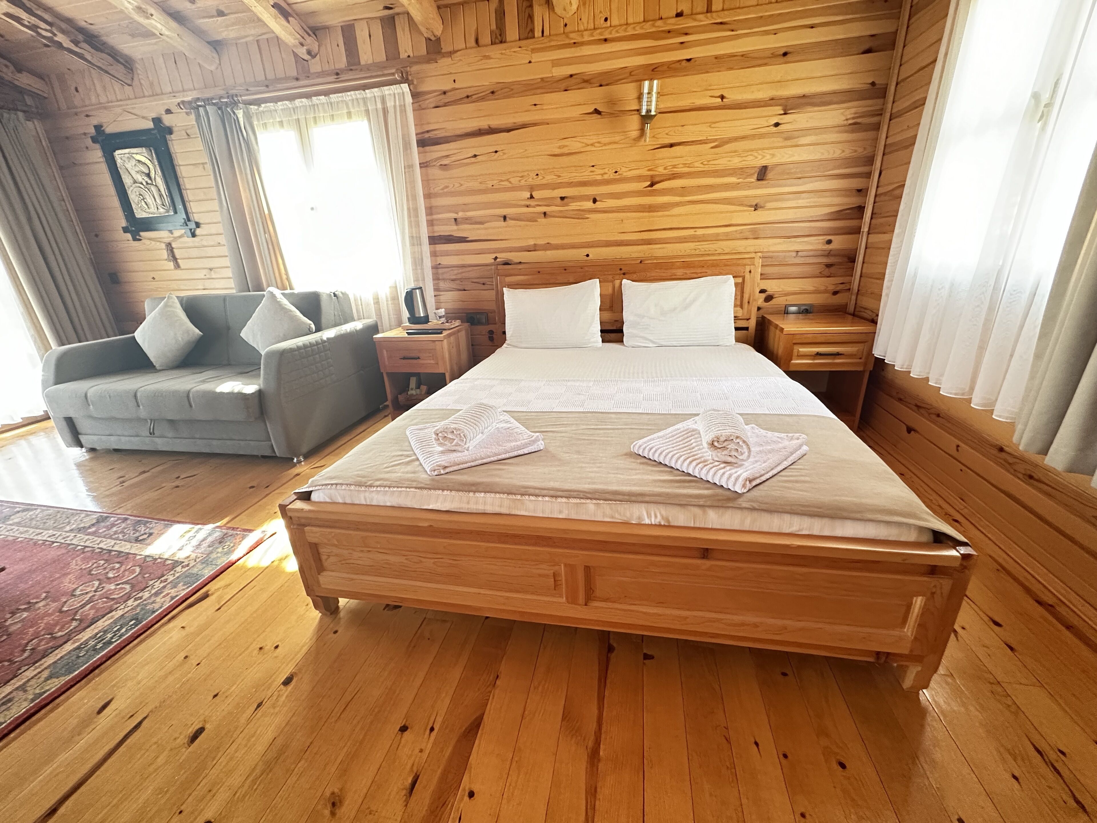 A wooden-paneled bedroom with a large bed, a grey sofa, and a nightstand with a lamp.