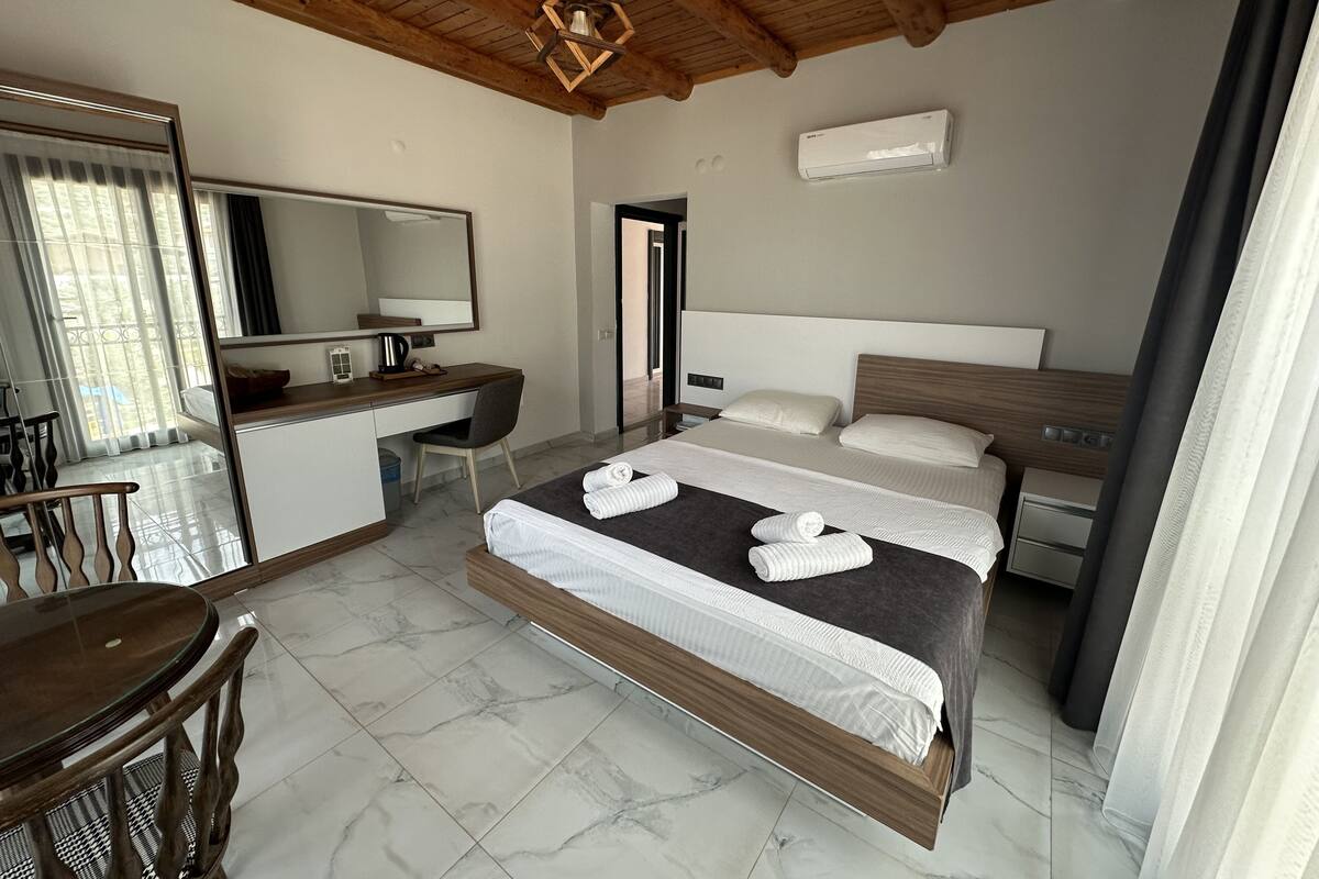 A hotel room with a bed, a desk, a chair, a mirror, and an air conditioning unit.