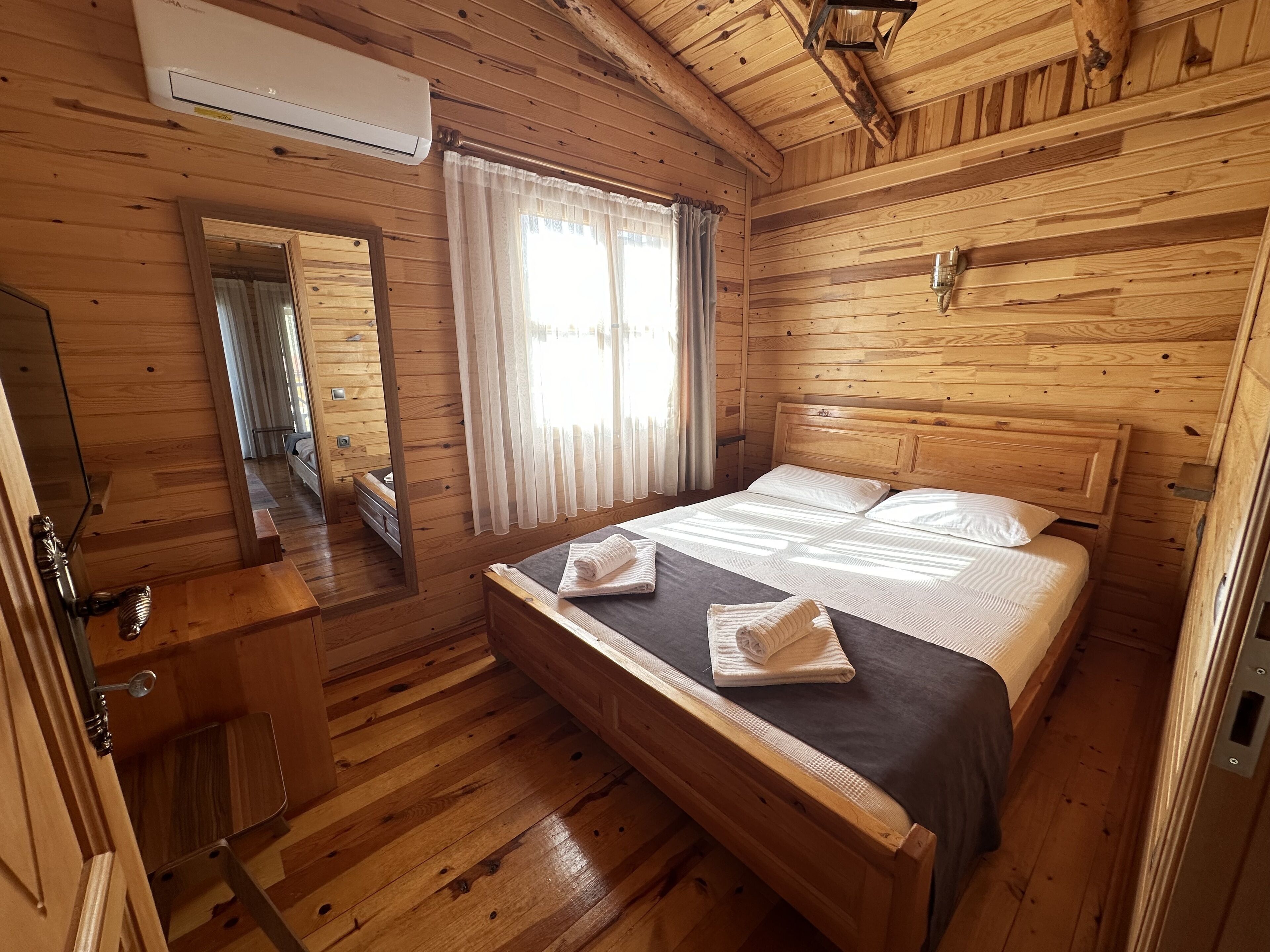 A wooden cabin room with a bed, a mirror, a window with curtains, and a wooden dresser.
