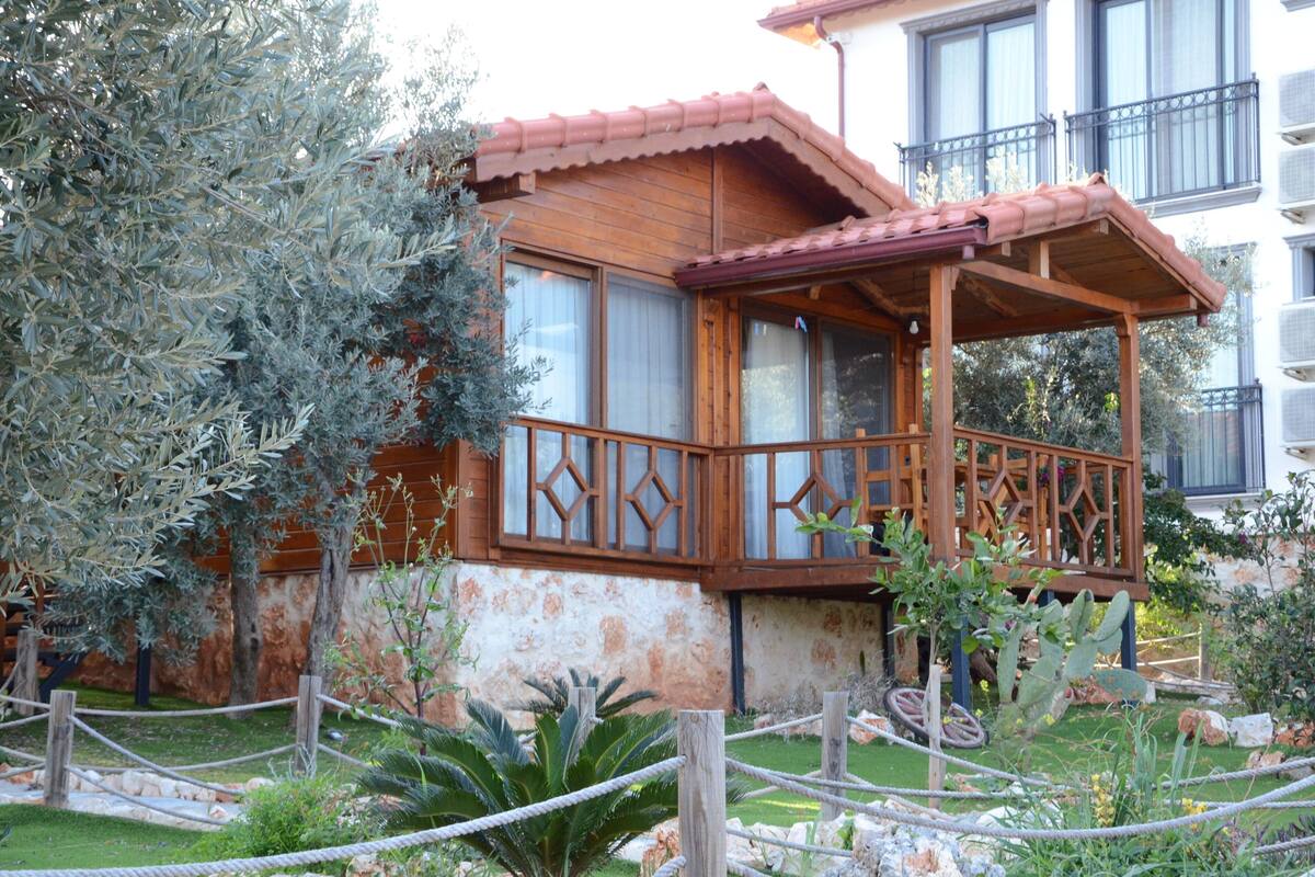 A wooden house with a balcony, surrounded by a garden with various plants and a fence.