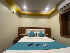 Habitación de hotel con cama, dos almohadas, colcha azul, dos controles remotos, mesita de noche y aire acondicionado.