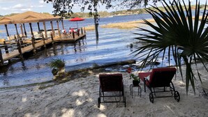 Ein Strandabschnitt mit Sand, zwei rote Liegestühle, ein kleiner Tisch und ein roter Sonnenschirm. Eine Holzstele führt ins Wasser, wo ein Boot und Sitzgelegenheiten zu sehen sind.
