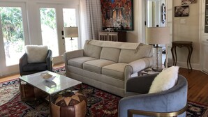Ein Wohnzimmer mit einem beigen Sofa, zwei Sesseln, einem Couchtisch und einem gemusterten Teppich. An der Wand hängt ein Gemälde und auf einem Beistelltisch steht eine Lampe.