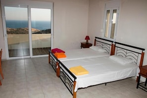 A bedroom with a bed, a nightstand, a lamp, and a sliding glass door leading to a balcony with a view of the sea.