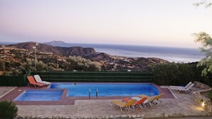 A swimming pool with two separate sections, surrounded by lounge chairs, set against a backdrop of hills and a view of the sea. - Paterakis Studio's Triopetra (Agios Vasileios)