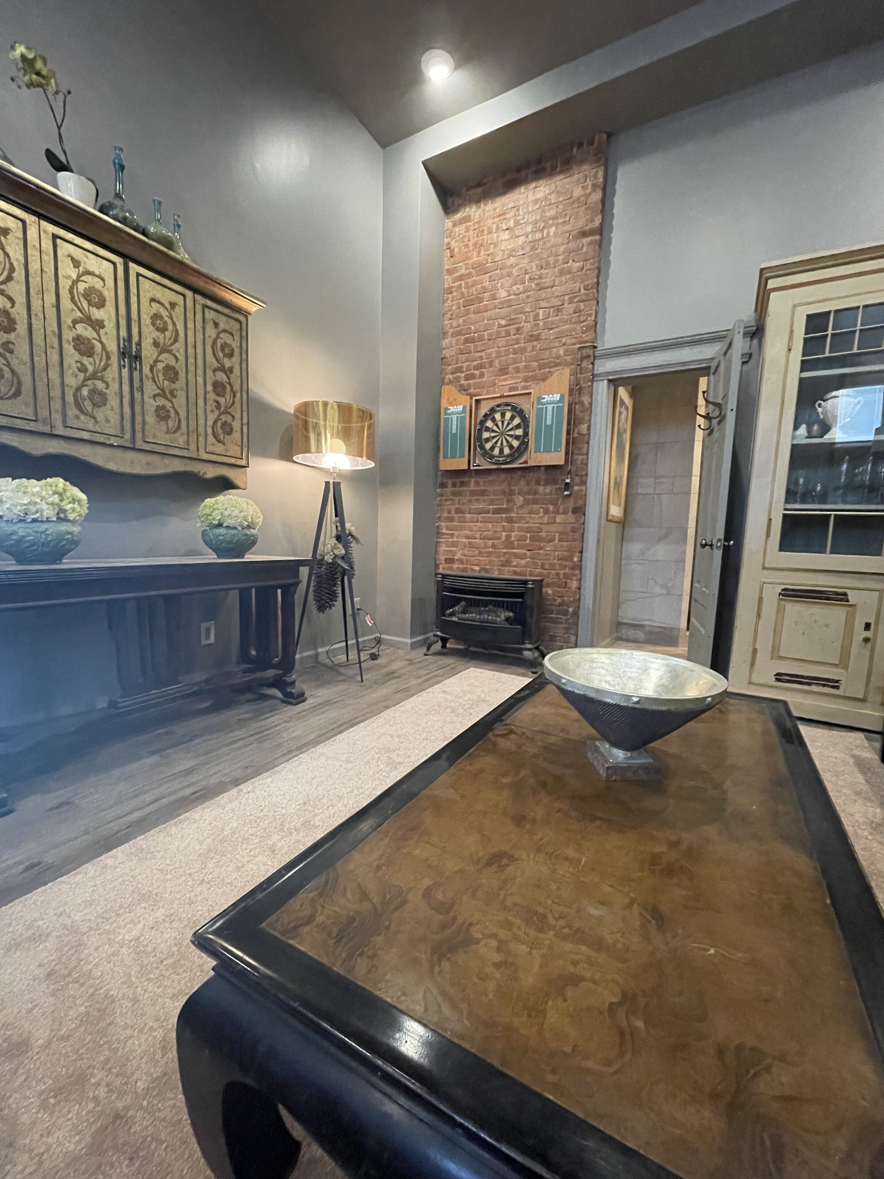 A room with a wooden coffee table, a fireplace, a dartboard, and a cabinet with decorative items.