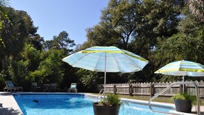 Una piscina con dos sombrillas, sillas de descanso y una valla de madera al fondo.