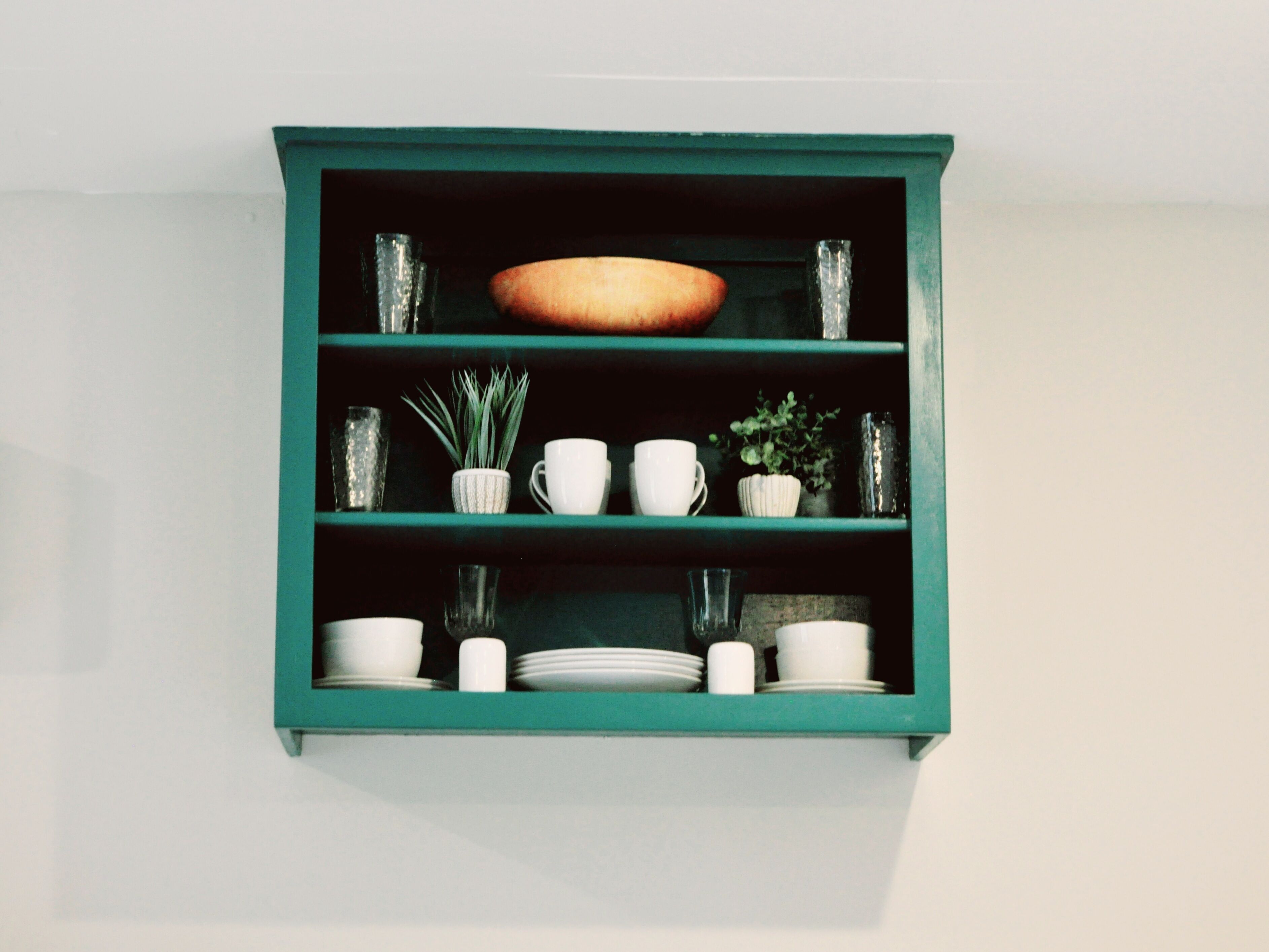 Um armário fixado na parede, com moldura azul-esverdeada e prateleiras que exibem diversos objetos de cerâmica branca, como xícaras, pratos e um pequeno vaso de planta.