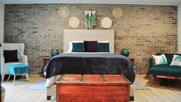 Un dormitorio con pared de ladrillo, cabecera acolchada, cama con ropa de cama oscura, un sillón azul, un sofá verde y un aparador de madera.