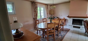 A dining room with a wooden table, chairs, a vase with flowers, a fireplace, and a window with curtains.