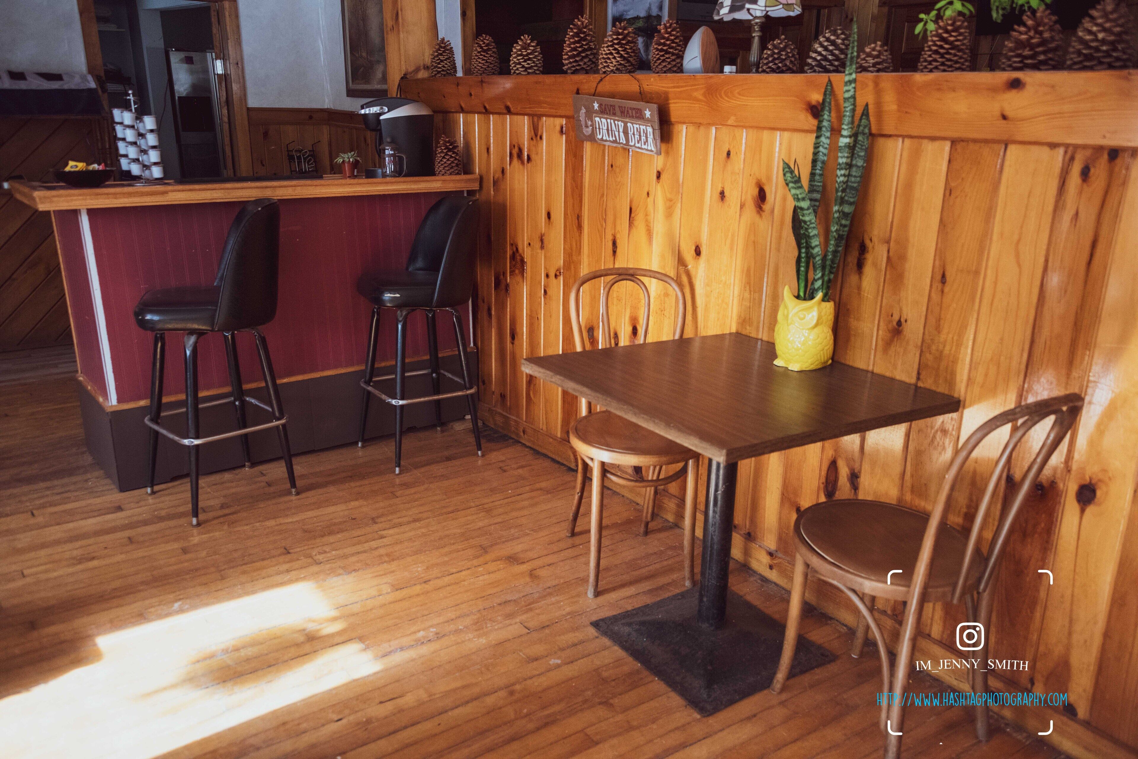 Una zona de bar con barra de madera, un cartel que dice "DRINK BEER", dos taburetes, una mesa y sillas.