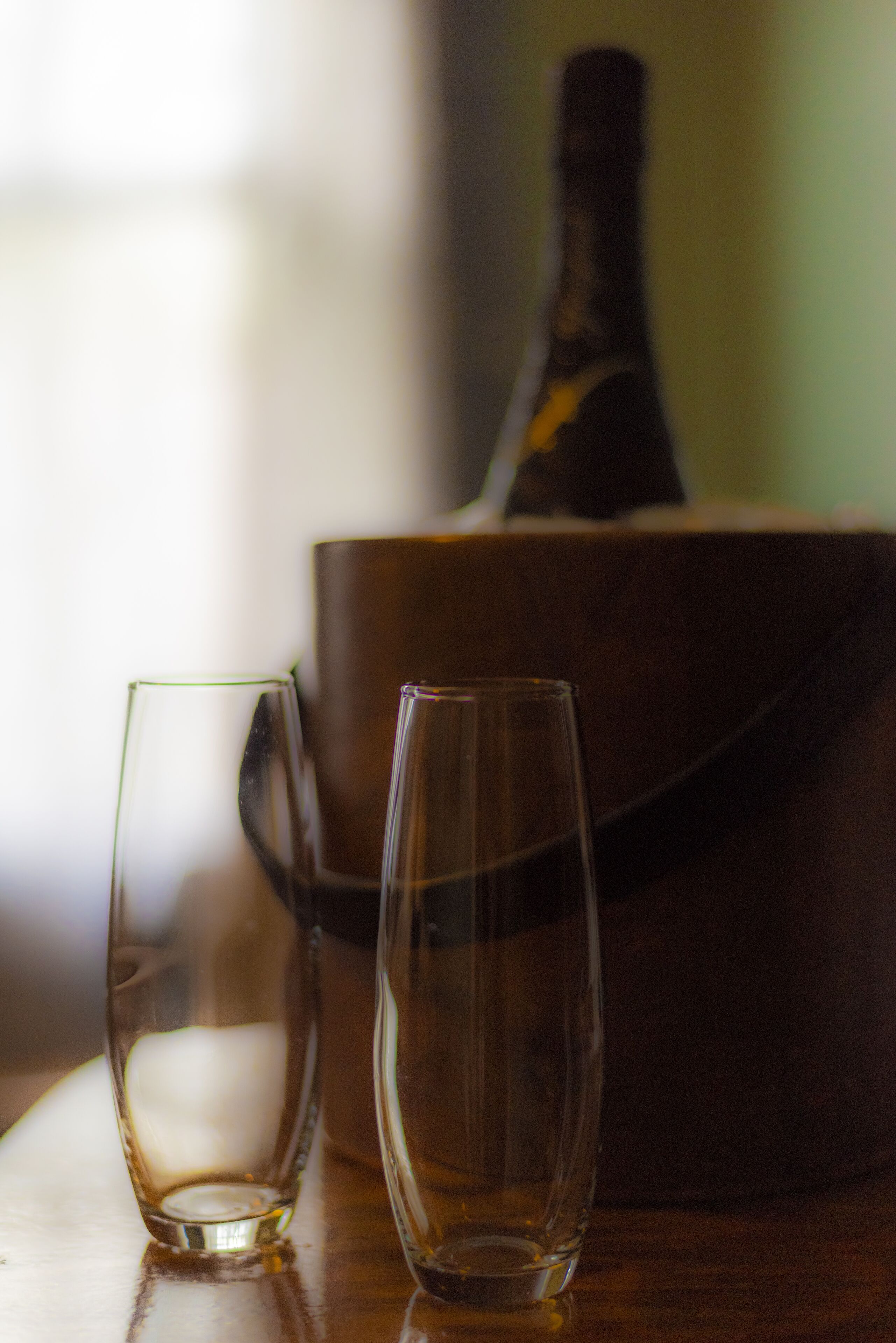 Two clear glasses on a wooden surface with a blurred bottle in the background.