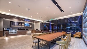 Um lobby de hotel moderno com uma longa mesa de conferência de madeira, assentos confortáveis, um balcão de recepção e um grande painel digital.