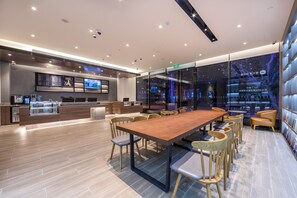 A modern hotel lobby with a long wooden conference table, comfortable seating, a reception desk, and a large digital display.