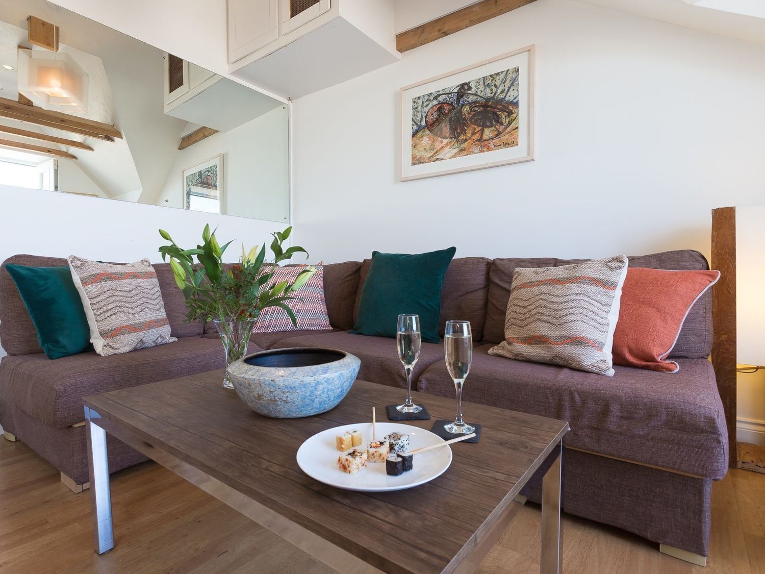 A modern living room with a brown sofa, a wooden coffee table, and a large blue bowl. There are two glasses of water and a plate with sushi on the table.