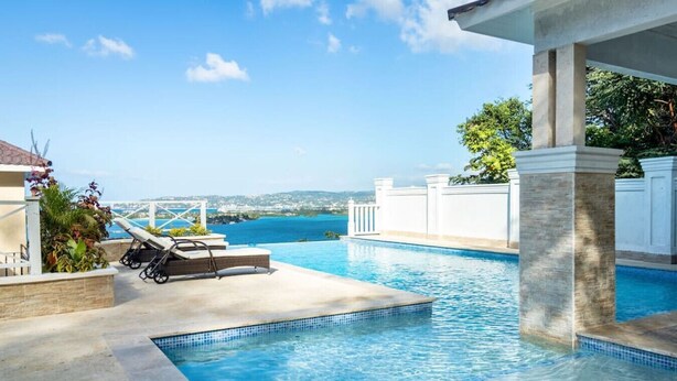 A swimming pool with a view of the sea, lounge chairs, and a white fence.