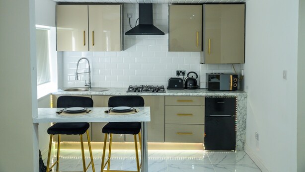 Une cuisine de taille réduite, équipée d’une cuisinière à gaz, d’un évier et d’un réfrigérateur compact. Un petit coin repas comprend deux chaises et une table.